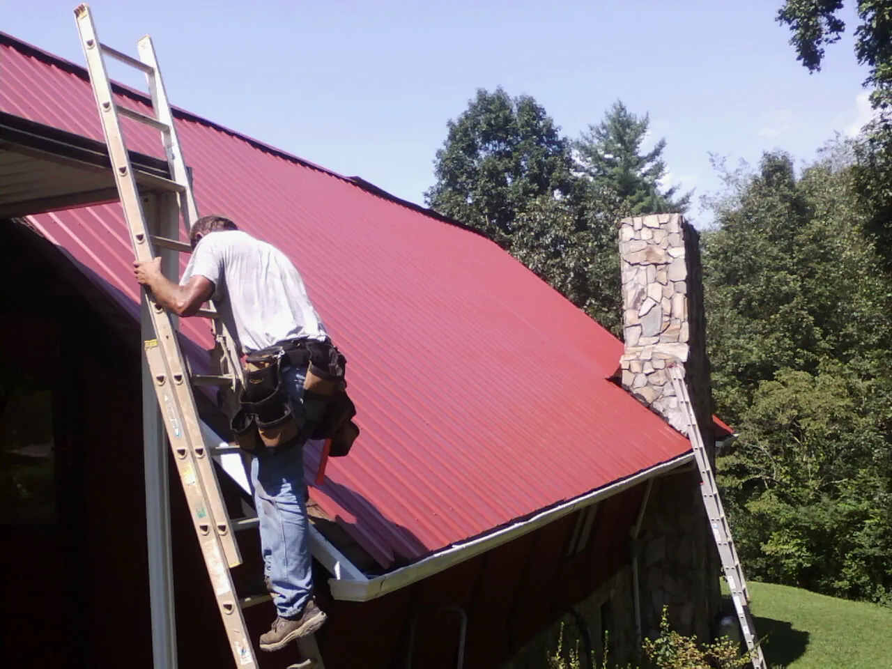 Roofer on a ladder beside a red metal roof and stone chimney — Soffit & Fascia Repair by Roofing Brothers in Wessington
